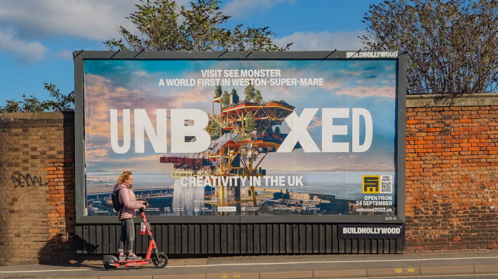 A person riding on a scooter past a large billboard promoting UNBOXED with the picture of the SEE MONSTER oil rig